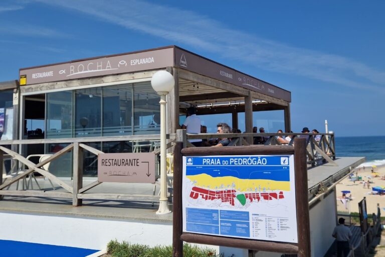 Restaurante A Rocha overlooks Praia do Pedrógão, with a beach map sign in the foreground and people dining under a shaded terrace.