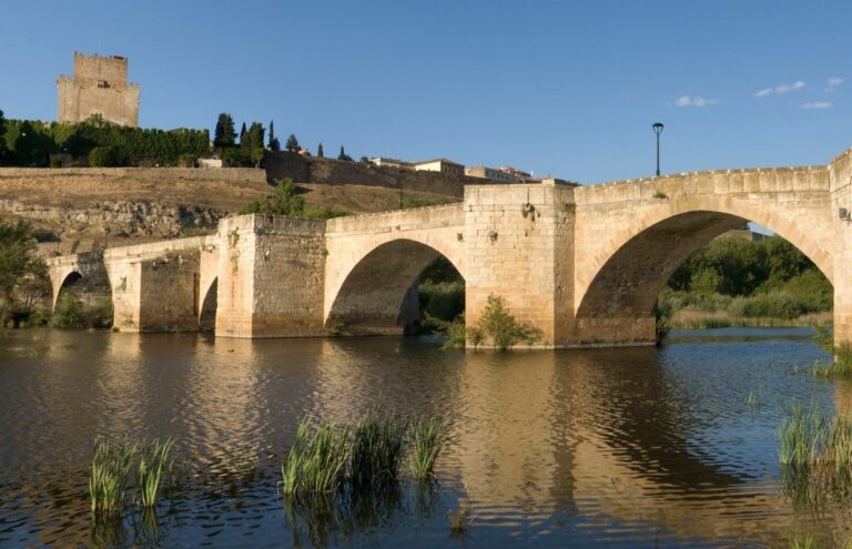 Historic stone bridge over a calm river with a castle in the background, set in the scenic landscape of Ciudad Rodrigo.