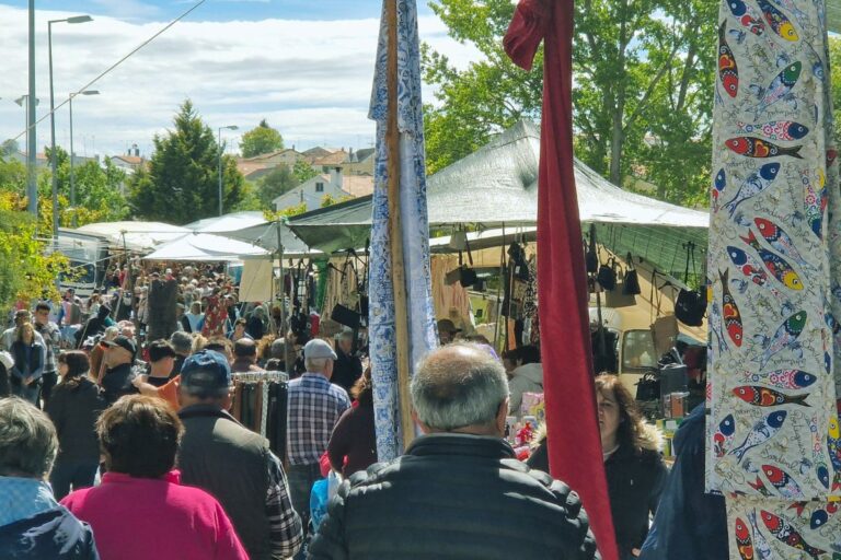 Crowds explore stalls under tents at the Feira de Vilar Formoso, with colorful fabrics and trees in the background.