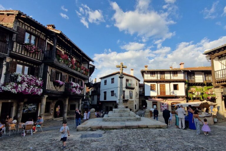 La Alberca Plaza Mayor features a stone cross, surrounded by traditional wooden buildings adorned with flowers and people strolling.
