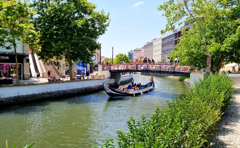 A Moliciero boat with passengers navigates a canal under a pedestrian bridge, surrounded by lush greenery and buildings on a sunny day.