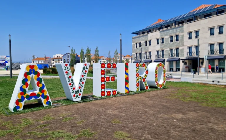 Colorful "Aveiro" sign with decorative patterns on a grassy area, modern building in the background under a clear blue sky.